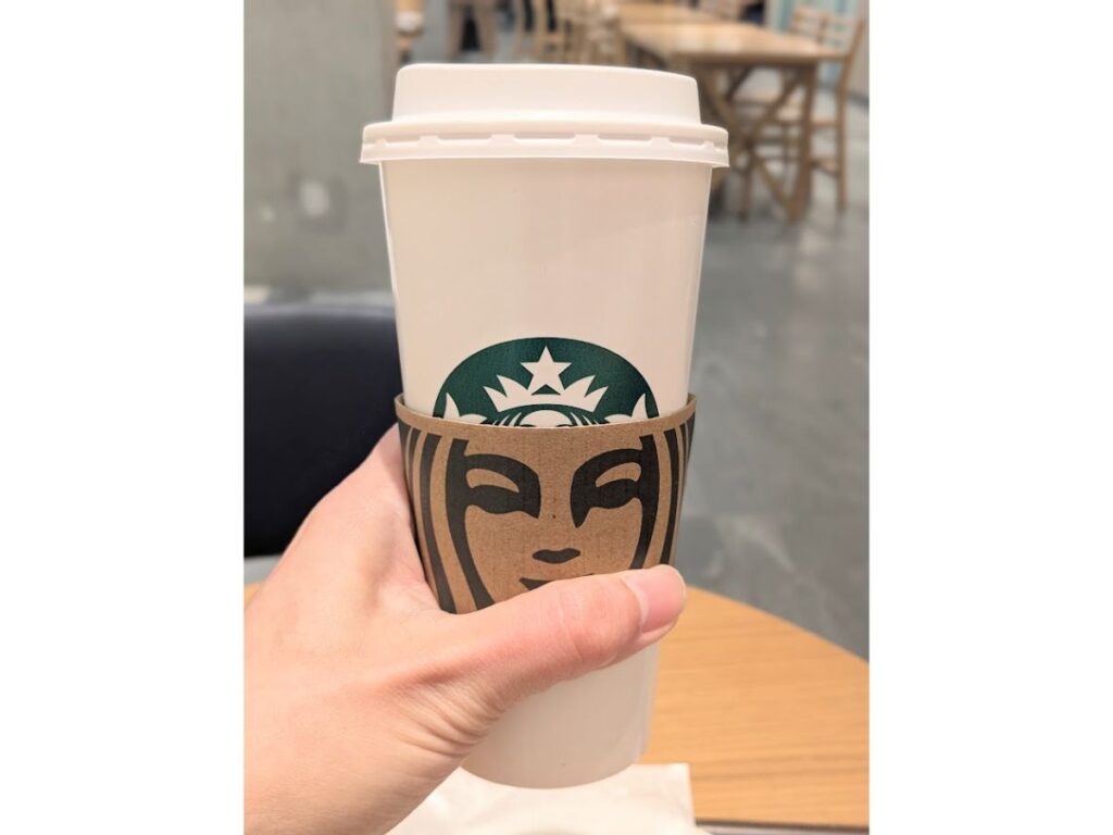 「スターバックスのベンティサイズの白い紙カップを手で持っている写真（店内）」